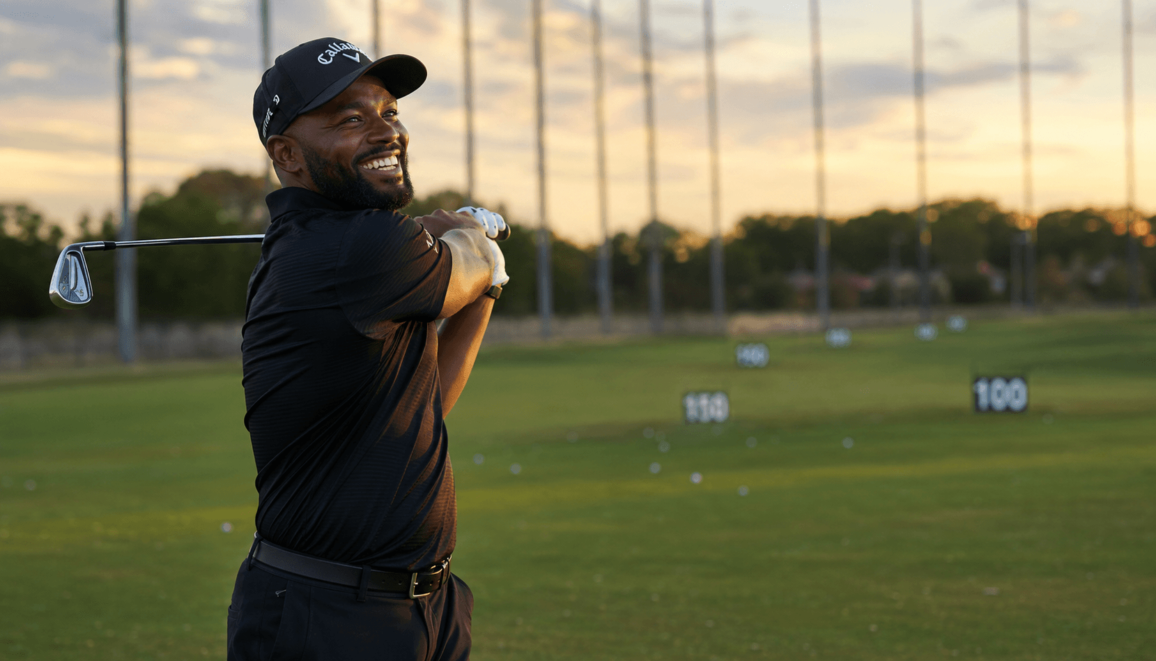 A golfer mid-swing on a practice range at sunset.
